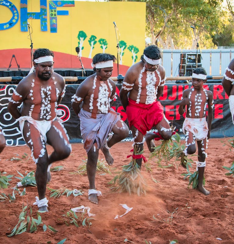 5 Indigenous men & 1 child dressed in traditional clothing & body paint dance in front of a stage.